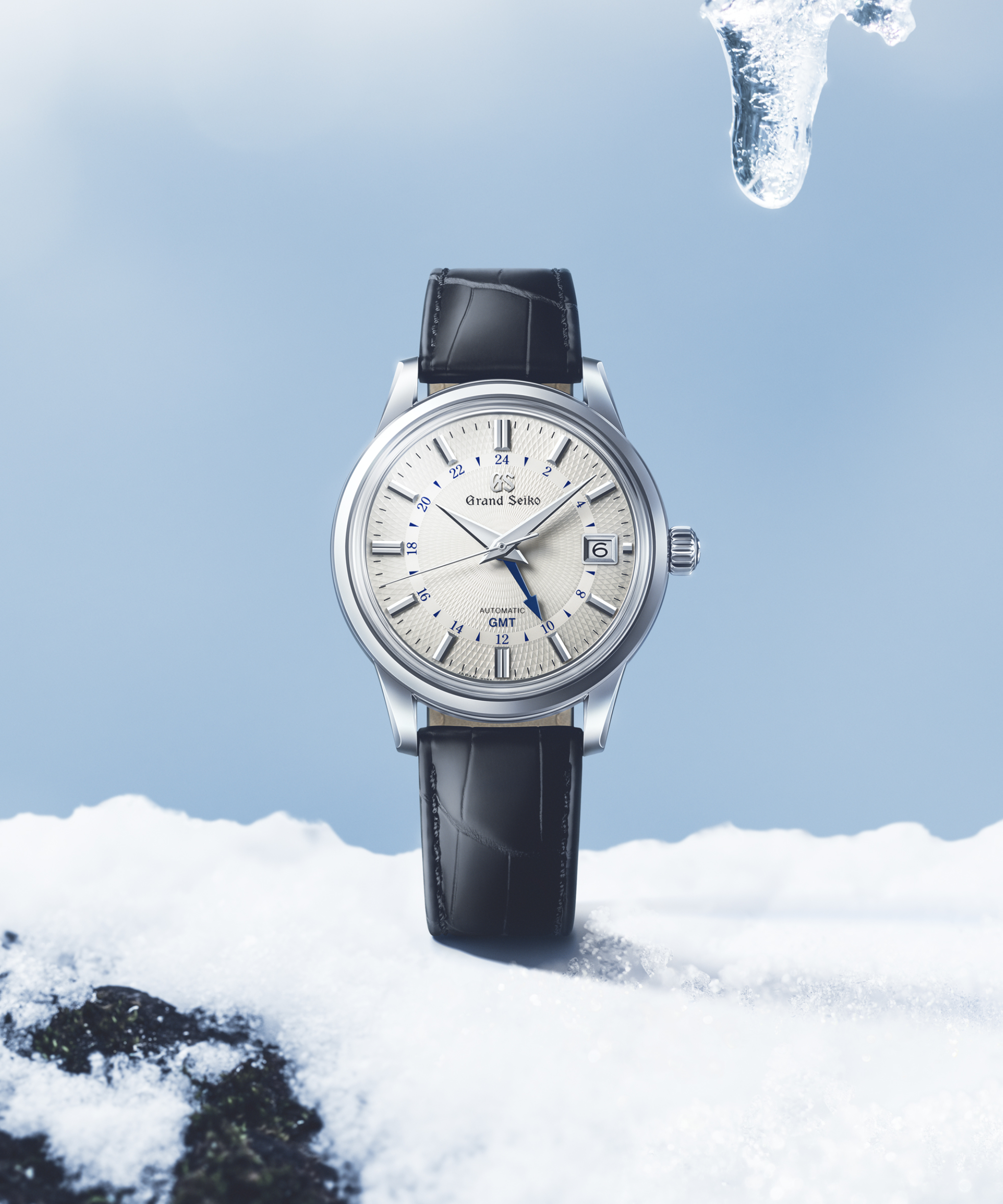 Reloj Grand Seiko SBGM255 Snowdrop con esfera blanca texturizada, aguja GMT azul y correa de piel sobre fondo ambiental.